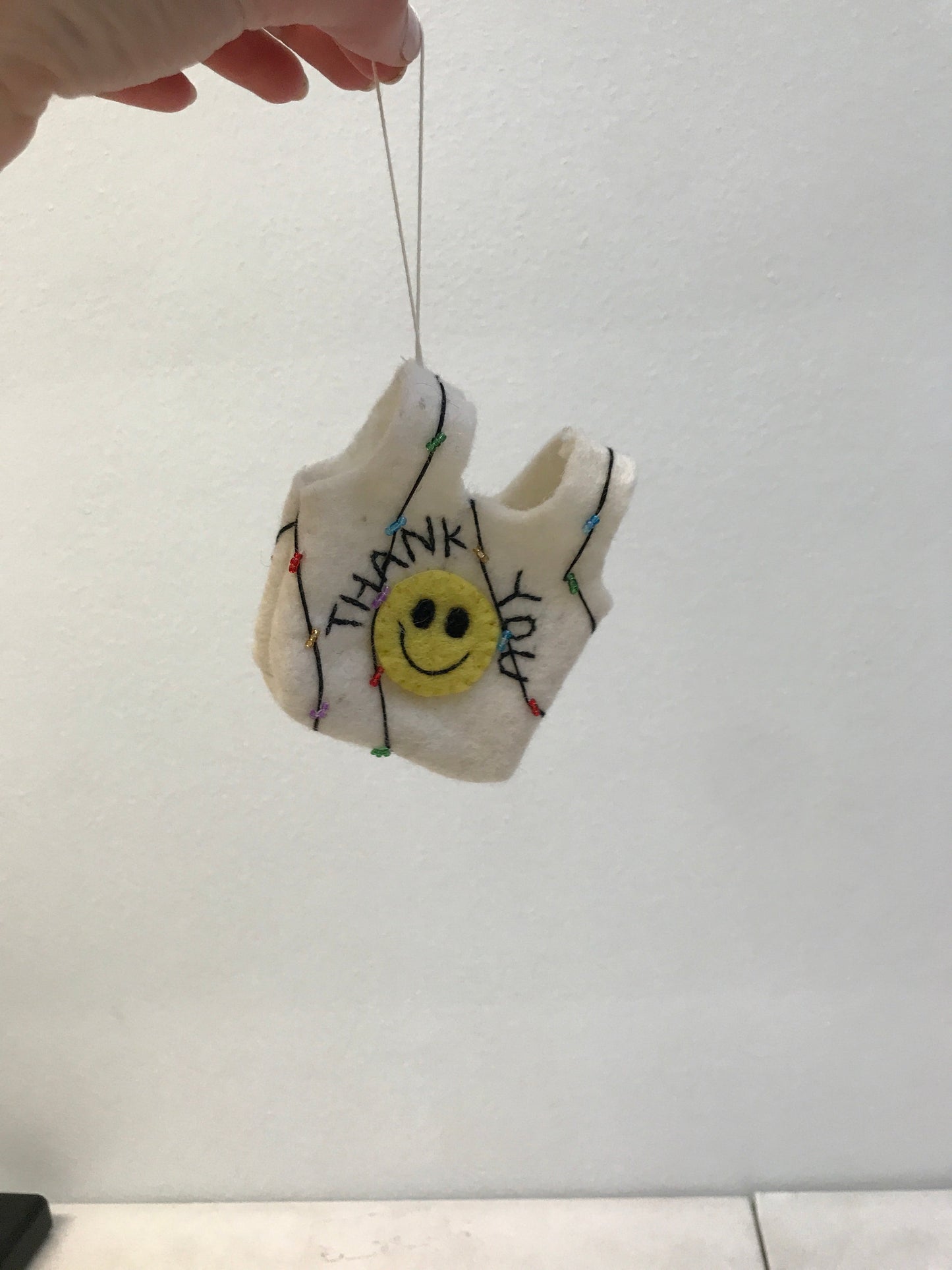 Felt ornament of a white thank you bag with yellow smiley face being held from the string by a hand.