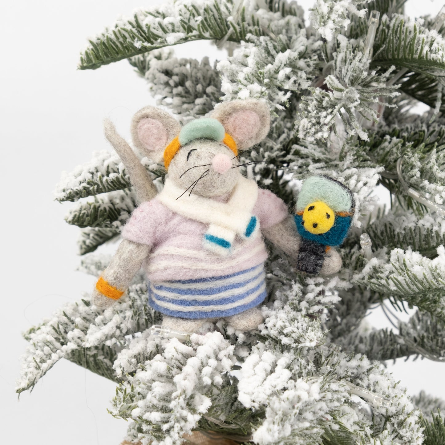 Felted ornament of a mouse dressed in a retro striped sweater, matching skirt, bright orange visor, and tiny sweatbands, with a pickleball paddle and ball in paw on a Christmas tree with fake snow.