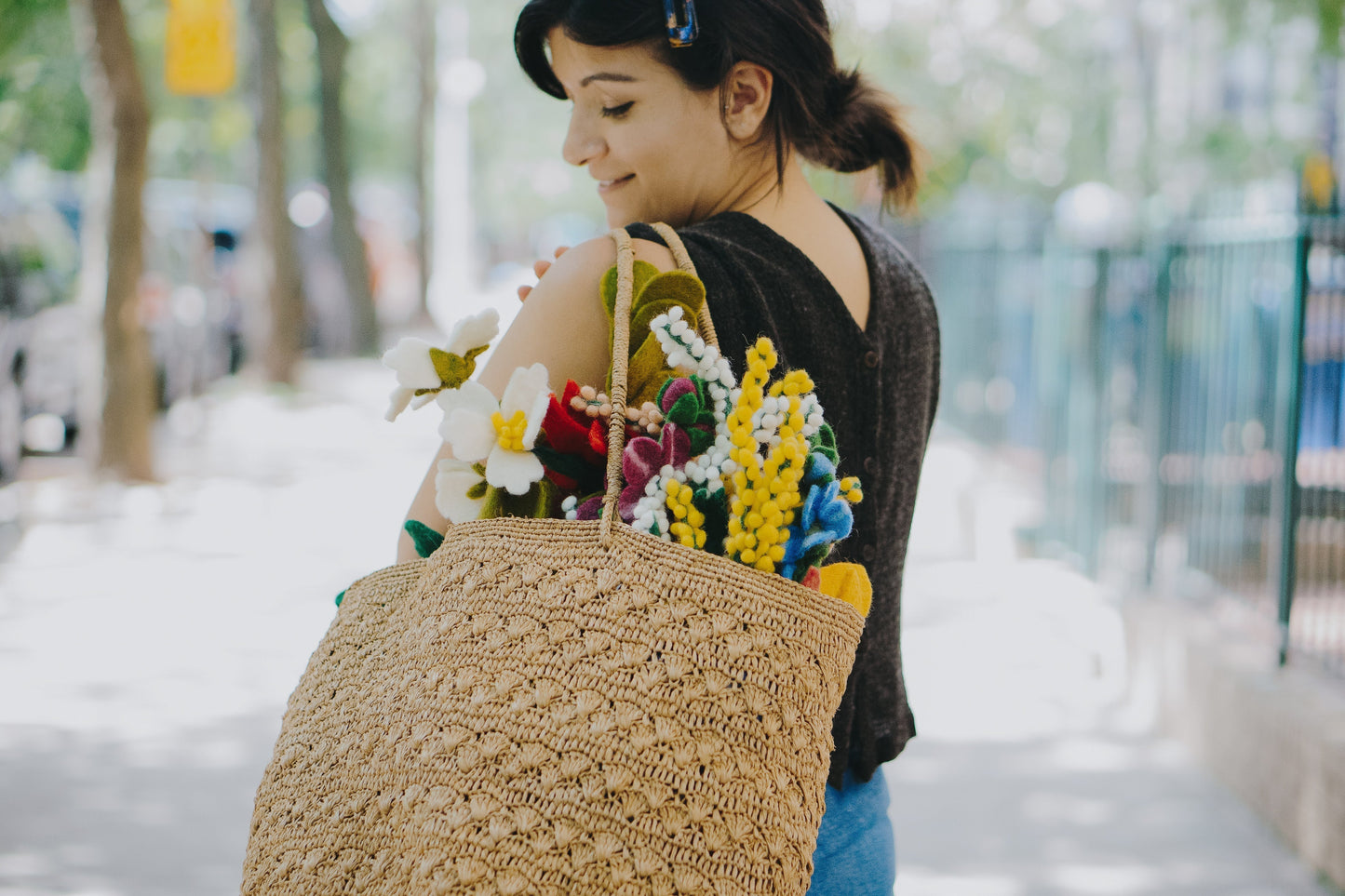 Bouquet of felt Baby's Breath flowers in peach, yellow, purple, white, and blue in tan woven bag on woman's shoulder.