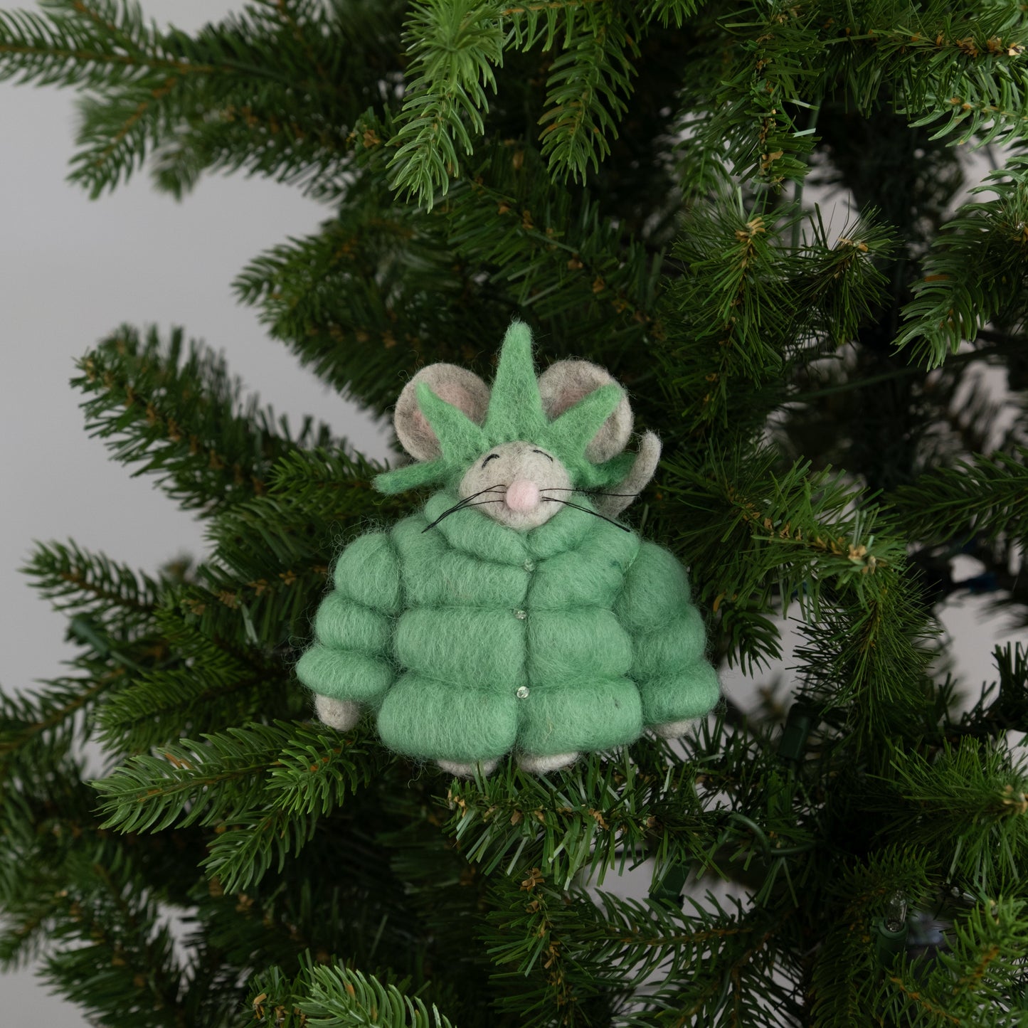Felt ornament of a mouse in a green puffer jacket and statue of liberty crown on a Christmas tree.