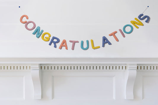 Felt garland spelling out Congratulations in pastel colors of peach, pink, teal, yellow, and blue hanging over a white mantel.