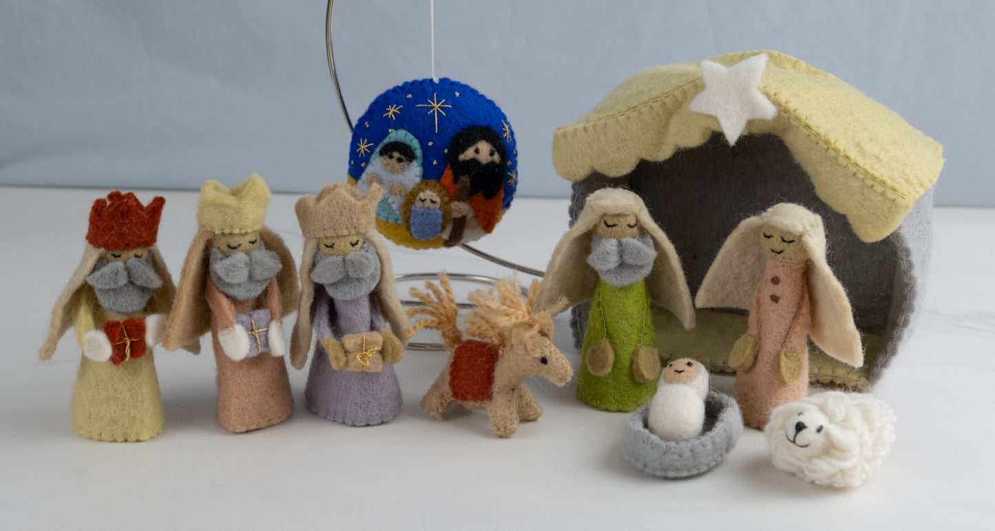 Felt ornament in a circle shape showing the nativity scene of Mary, Joseph, and baby Jesus underneath the night sky behind a full nativity scene set.