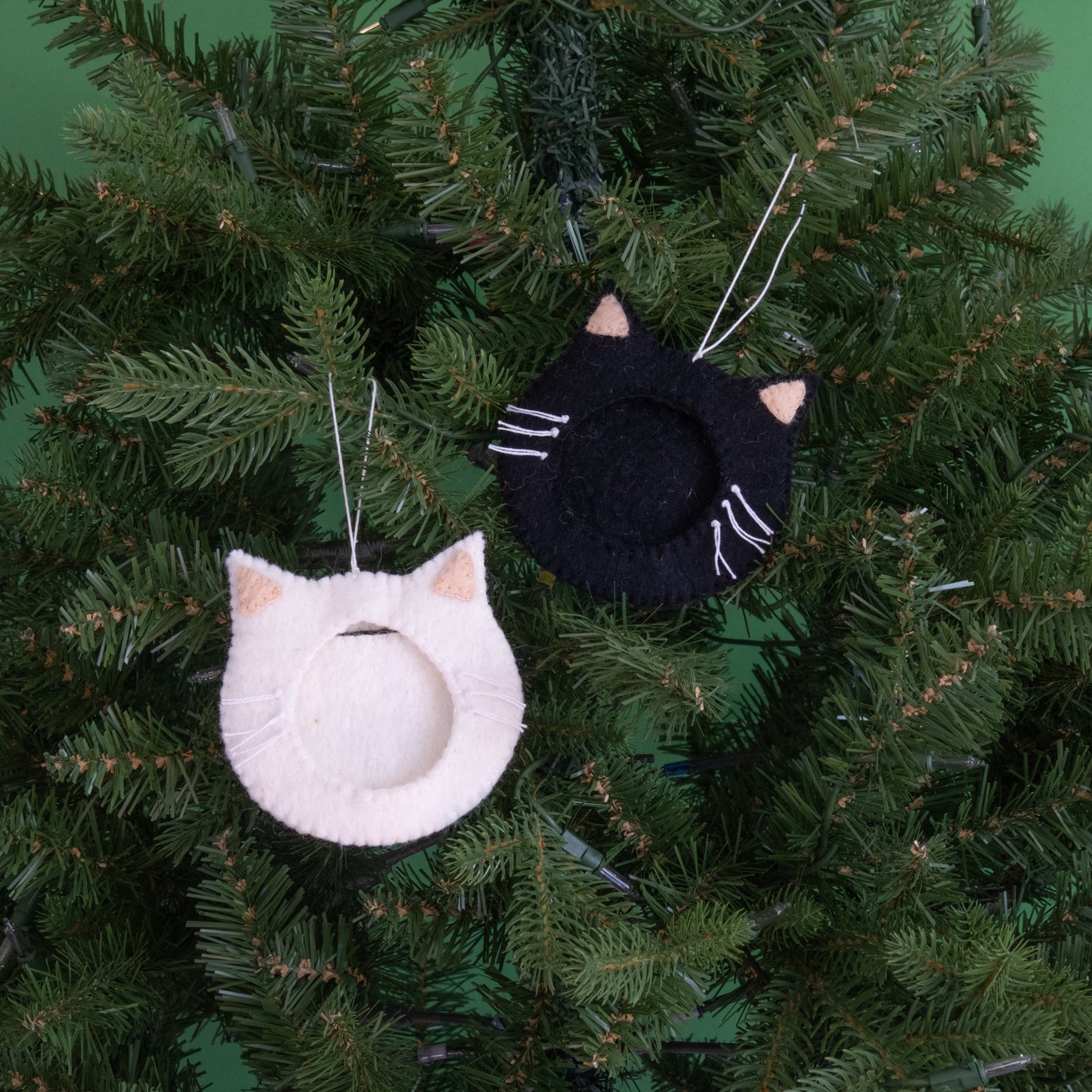 Felt ornaments of one black and one white cat face with a frame in the middle to put a photograph hanging on a Christmas tree.