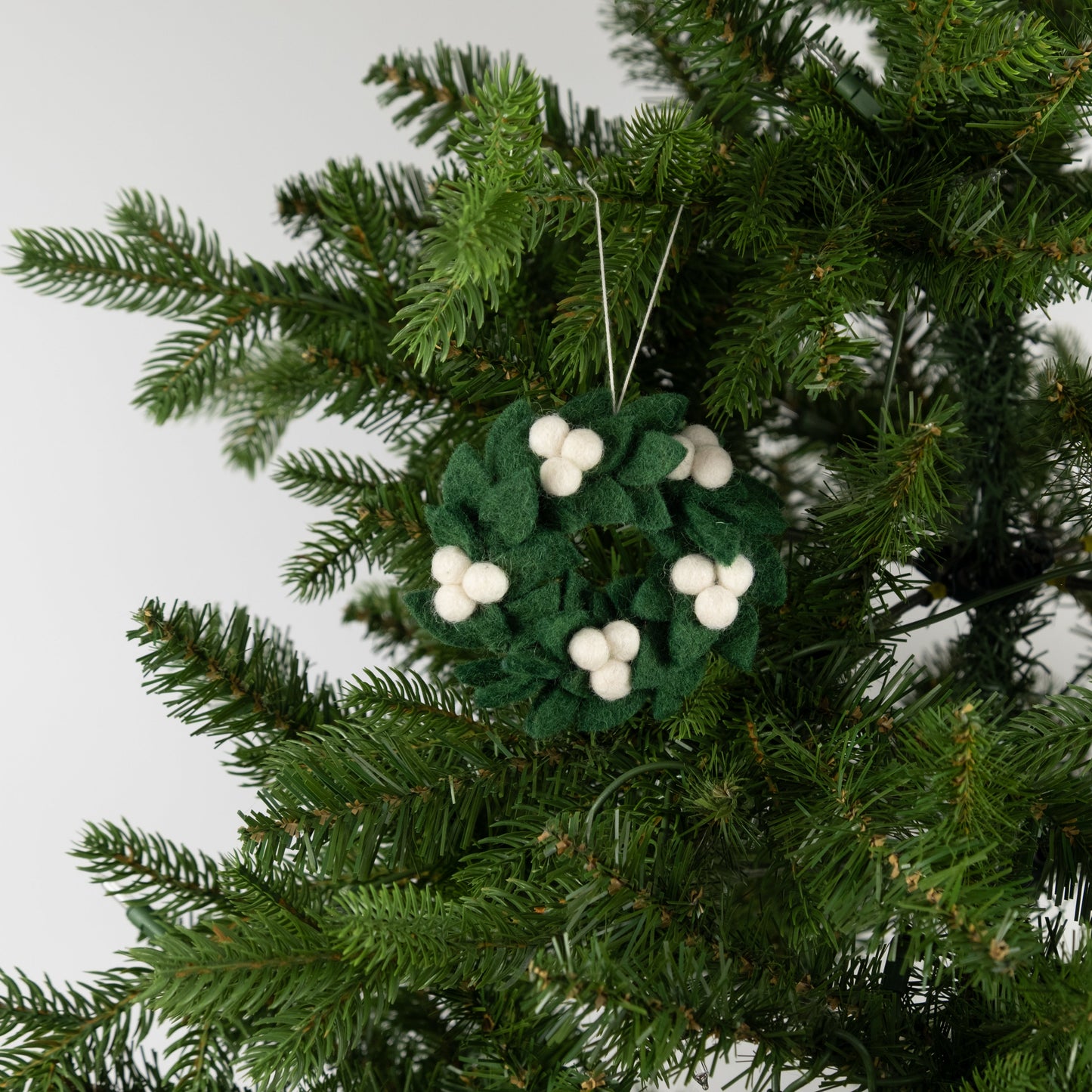 Felt ornament of a mini wreath in green with white berries hanging on a Christmas tree.