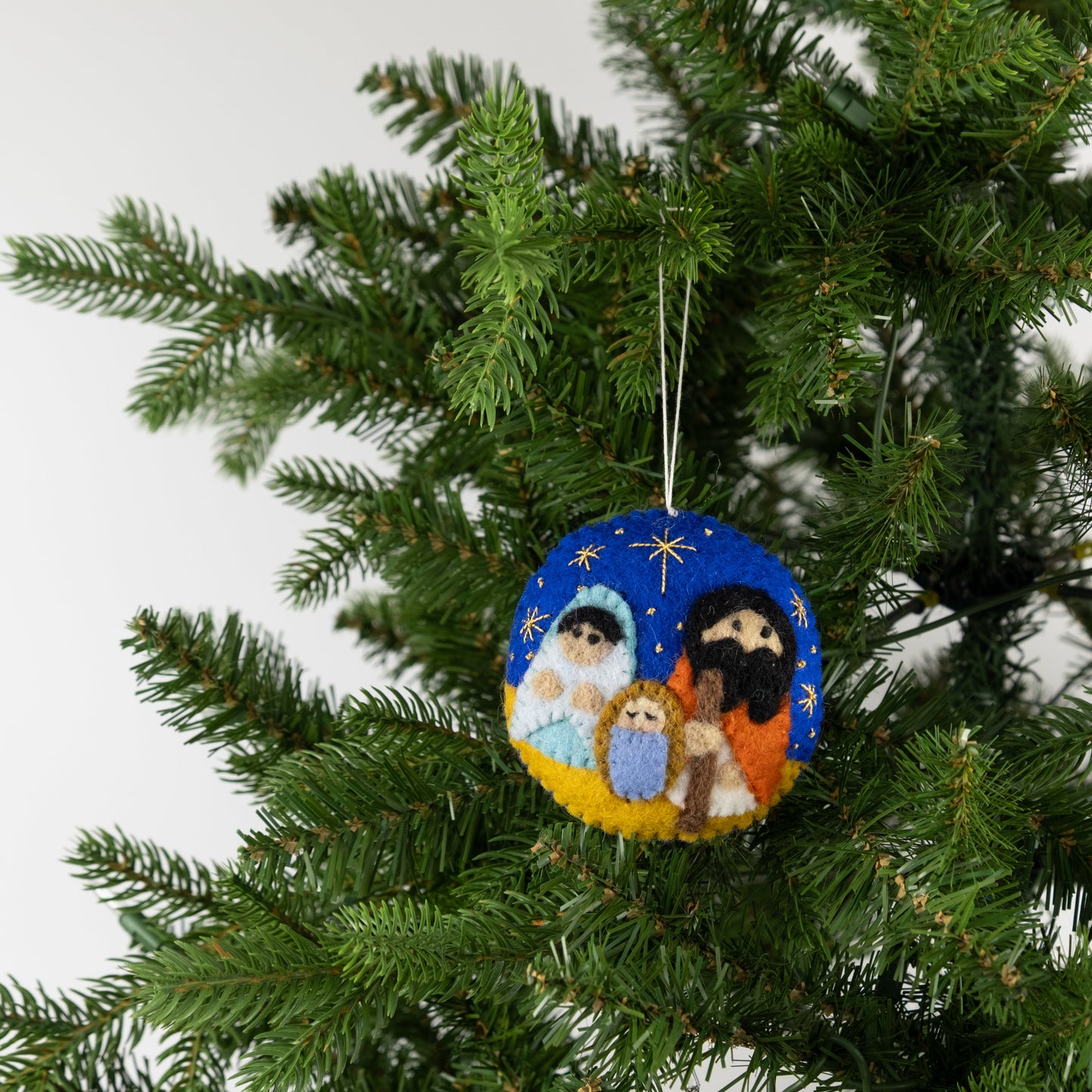 Felt ornament in a circle shape showing the nativity scene of Mary, Joseph, and baby Jesus underneath the night sky hanging on a tree.