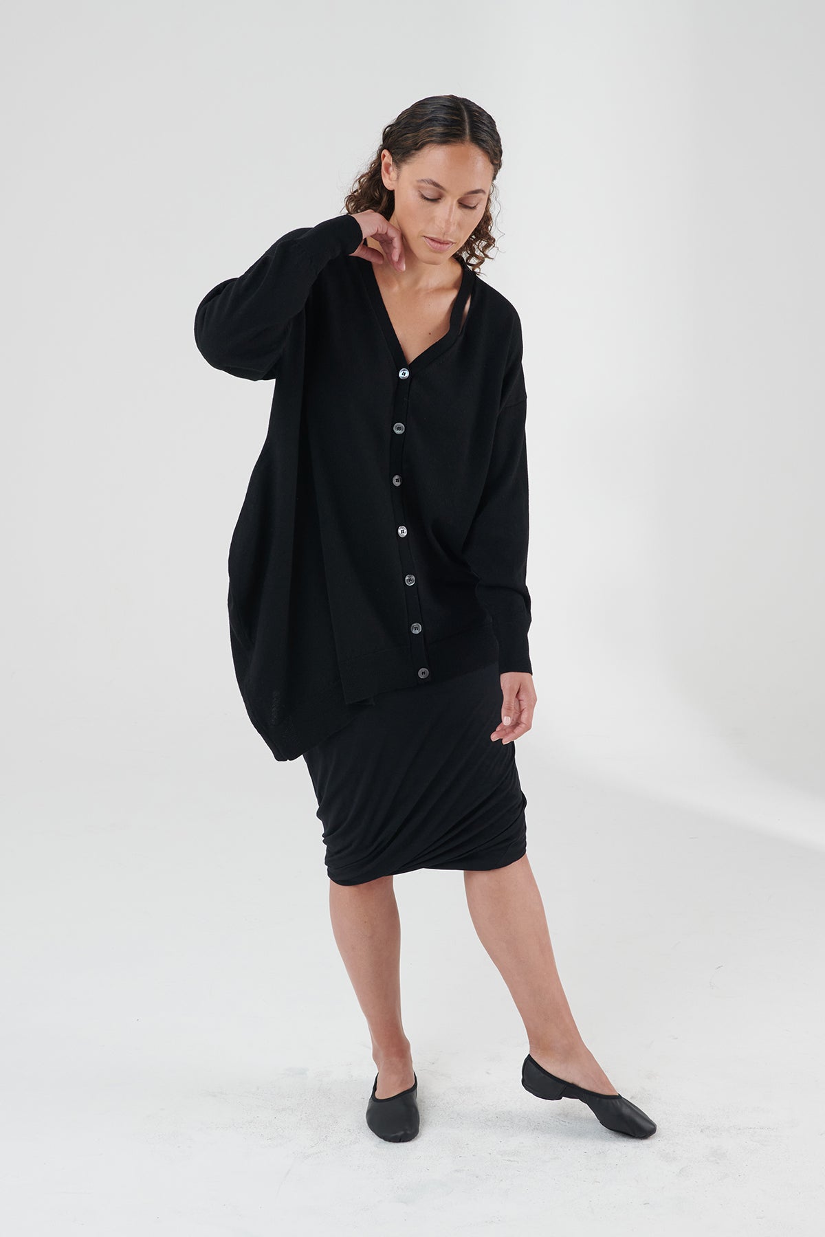 Woman wearing black cardigan with black skirt and shoes, looking towards the ground.
