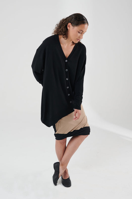 Woman wearing black cardigan with tan skirt and black shoes, looking towards the ground.
