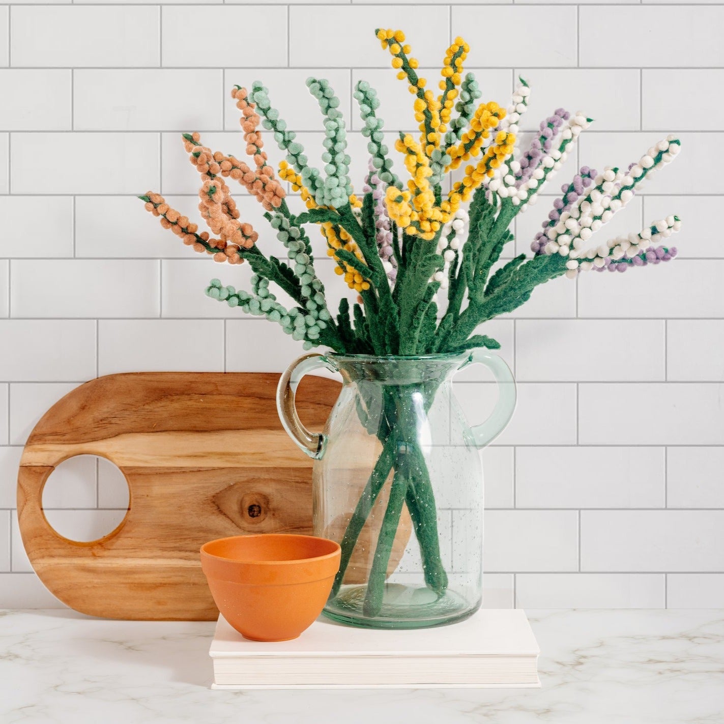 Bouquet of felt Baby's Breath flowers in peach, yellow, purple, white, and blue in glass vase on countertop.