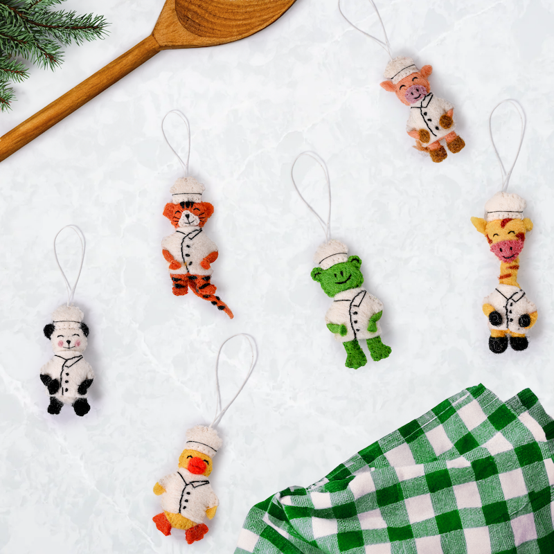 Scattered felt ornaments of animals in Chef's hats and coats showing a panda, duck, giraffe, frog, pig, and tiger on table with cooking spoon and green and white checkered kitchen towel.