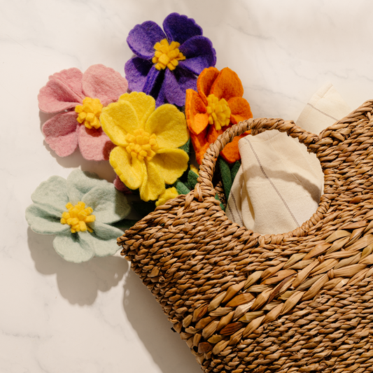 Bunch of felt Cosmo flowers in light blue, purple, yellow, orange, and pink in natural woven bag.
