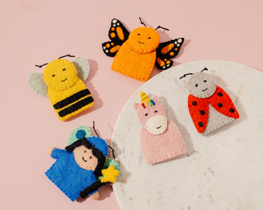 Scattered finger puppets of a pink unicorn, a blue fairy, an orange Monarch Butterfly, a Ladybug and a yellow Bumble Bee on a white and pink surface.
