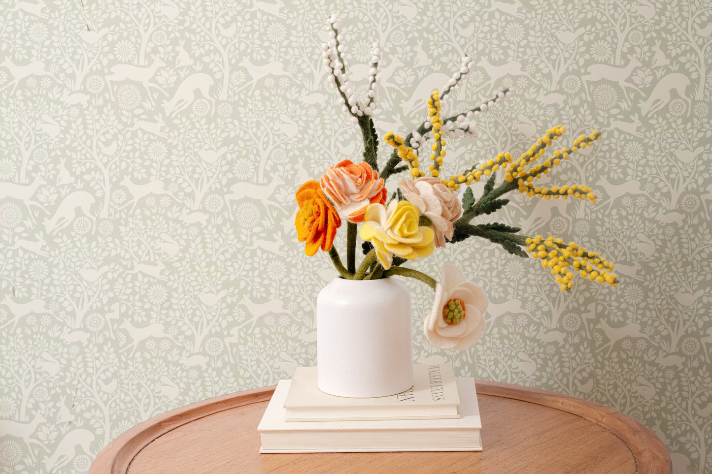 Felt floral arrangement in a white vase on a wooden table with a patterned wall background