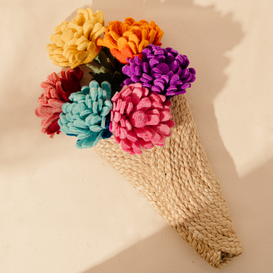 Bunch of felt flowers with many petals in yellow, orange, bright pink, purple, blue, and dusty pink in a woven natural cone hanging on wall.