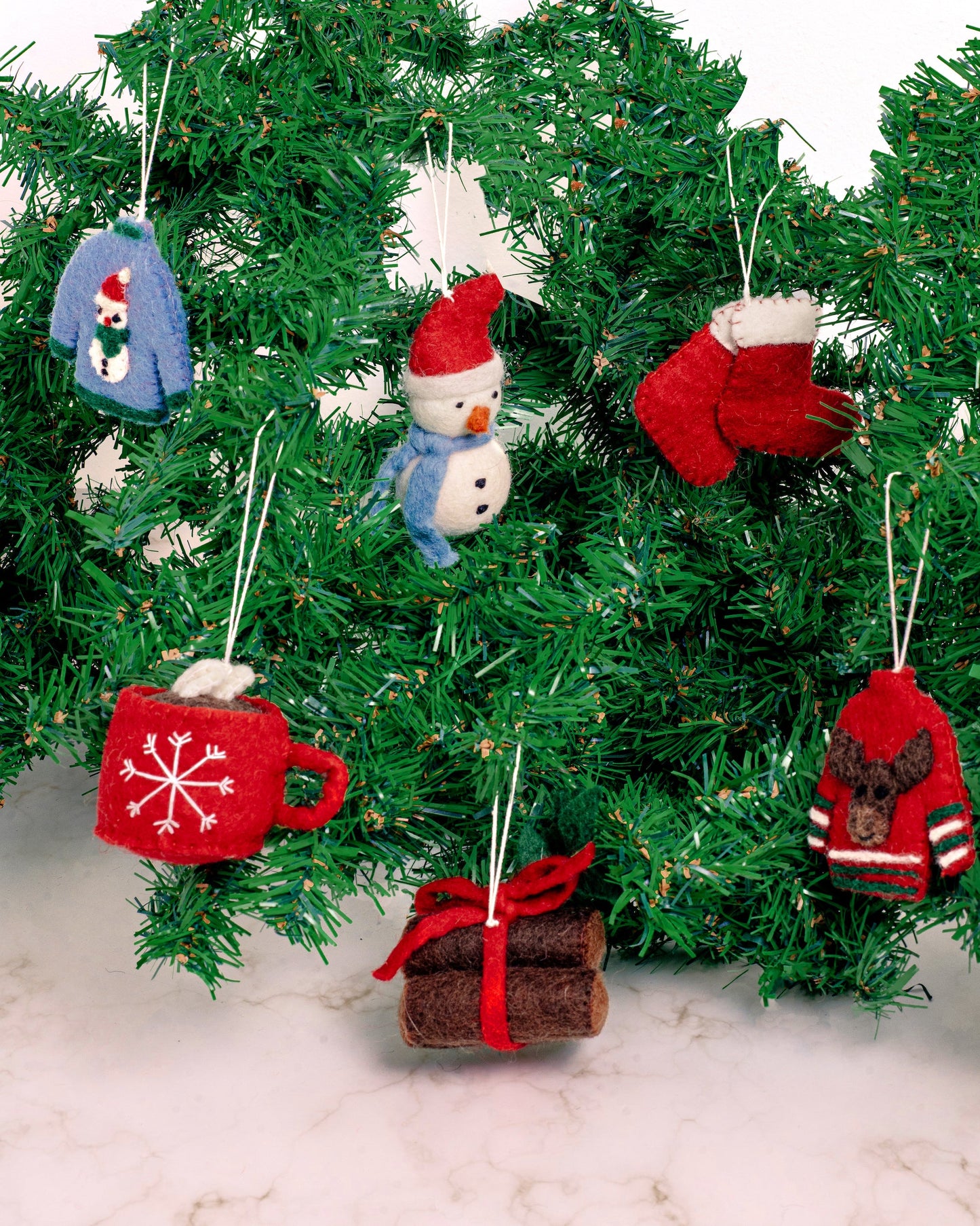 Felt ornaments of two red Christmas stockings, yule logs tied together with red bow, hot chocolate in red mug, grey sweater with snowman, snowman with red hat, and red sweater with a moose on it hanging on a Christmas tree.