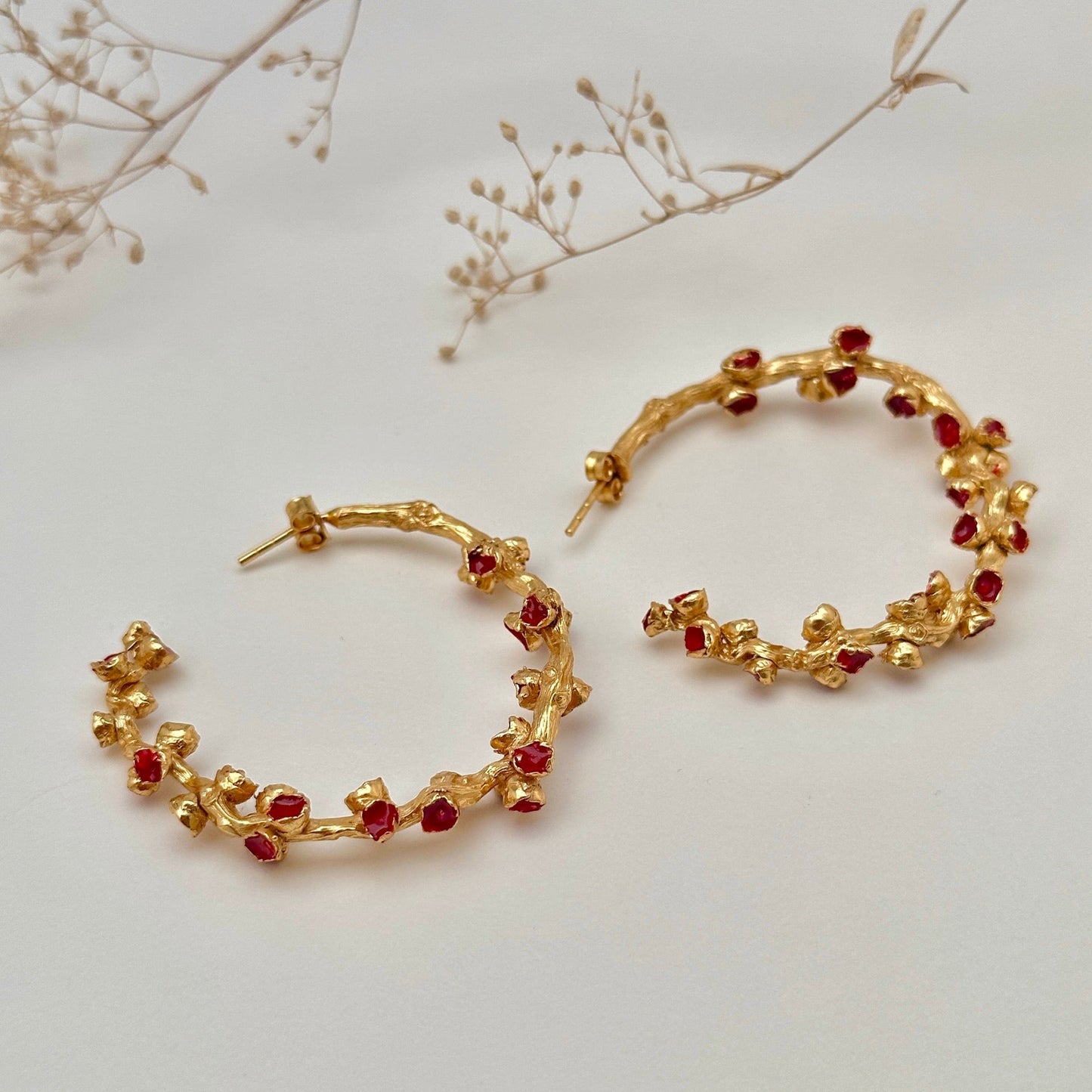 Pair of red and gold hoop earrings laying on side. Small dried flowers above.
