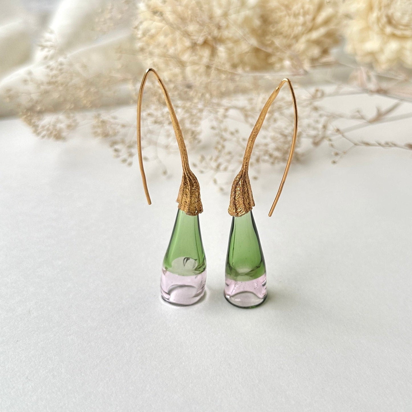 Pair of green and pale pink glass earrings with gold colored findings on grey background. Small dried flowers above image.