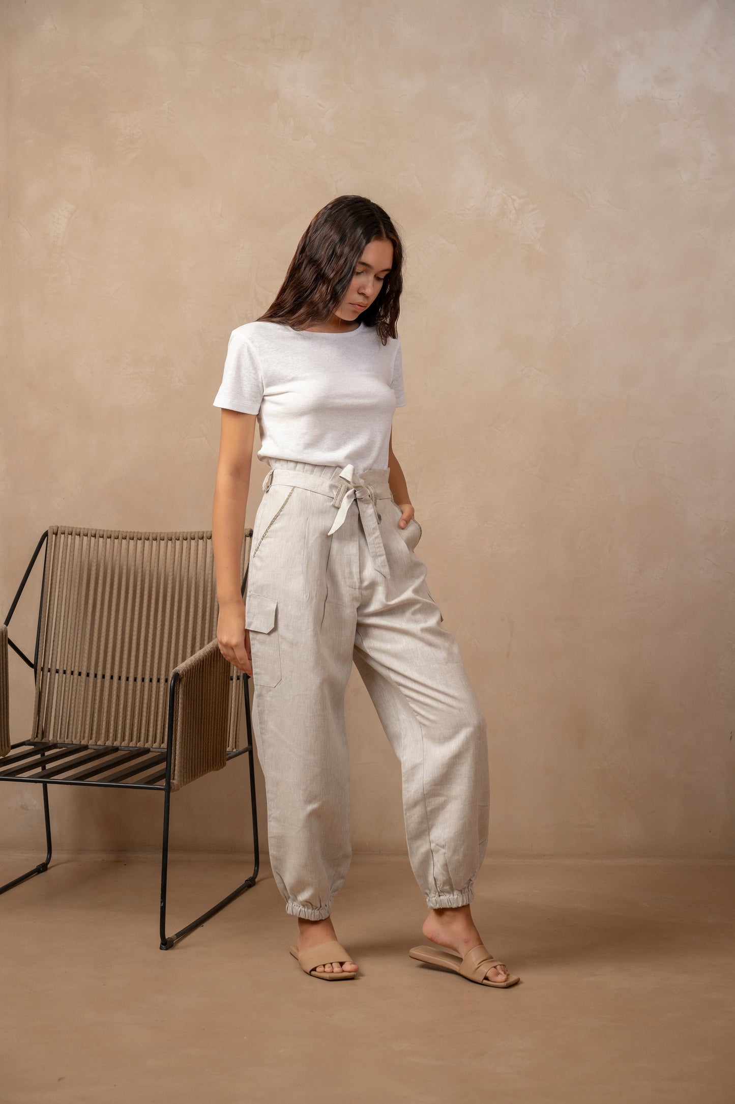 Woman wearing natural color Yama Cargo Jogger Pants, right hand holding onto black chair while looking at ground.