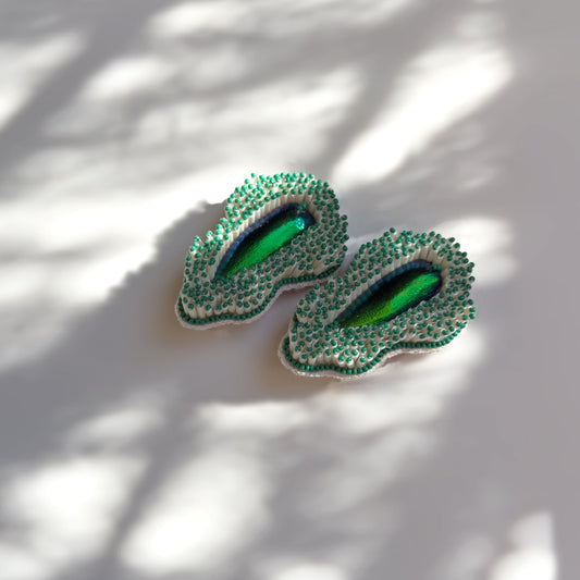 Pair of Pennata beaded green glass earrings with iridescent centers on shadowy background.