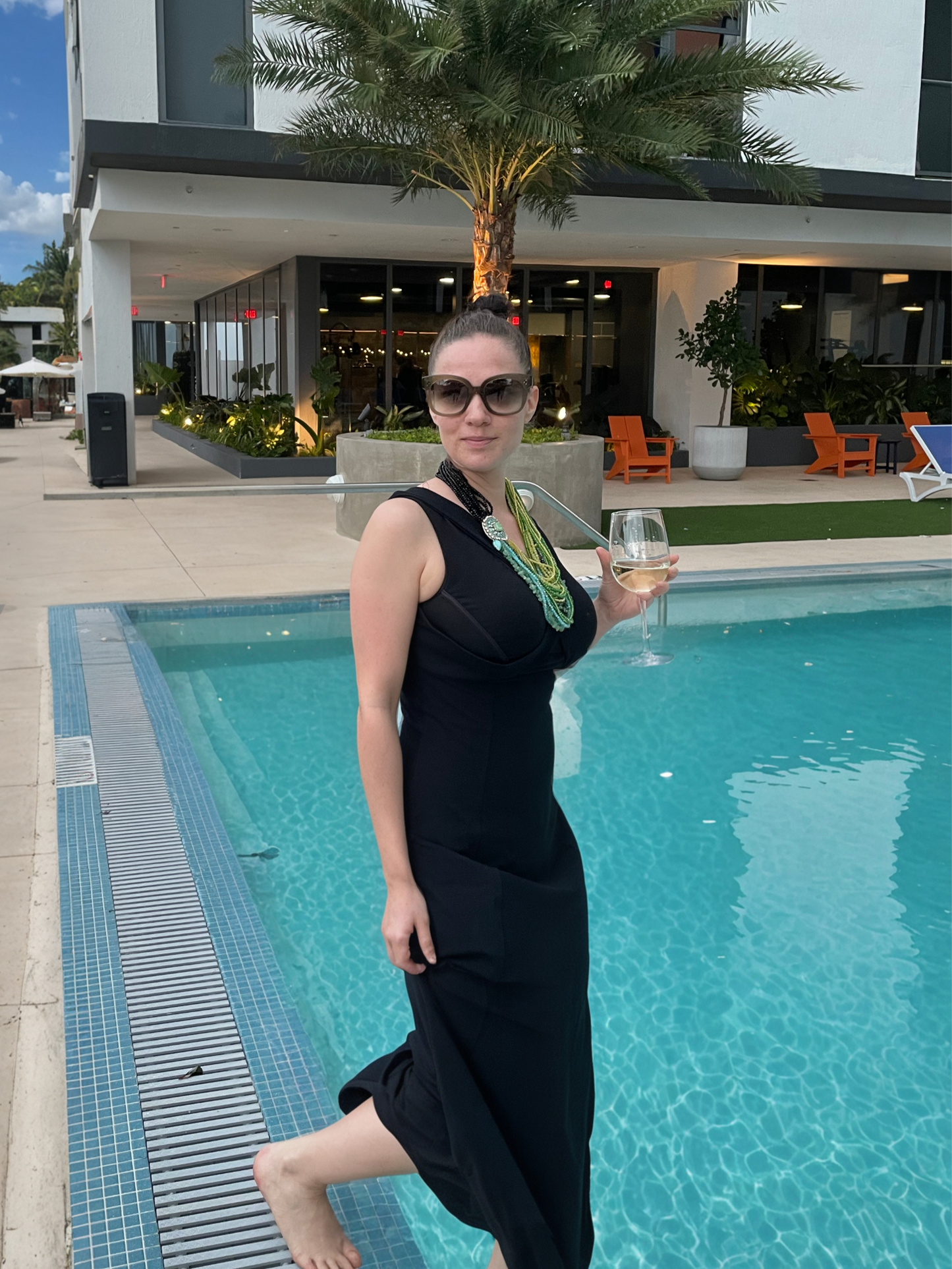 Woman wearing long black dress, holding a glass in left hand, stepping into a pool.