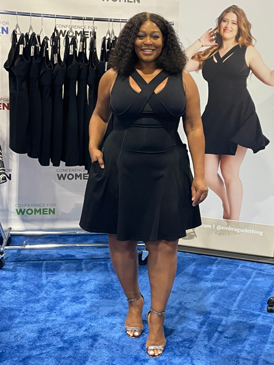 Woman standing on blue carpet, right hand in pocket, wearing black flare dress.