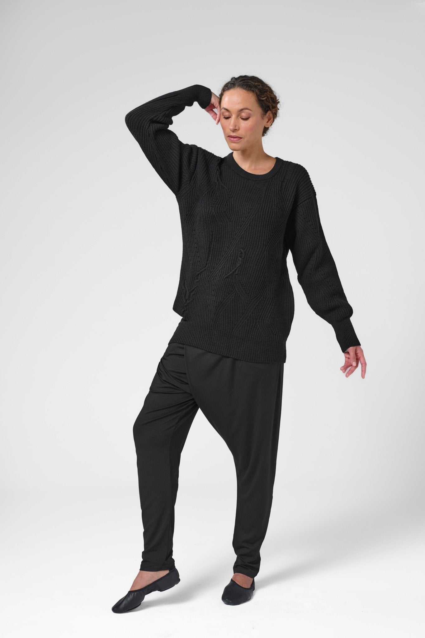 Woman with right elbow up wearing black crewneck sweater with asymmetric ribbed design and black pants.