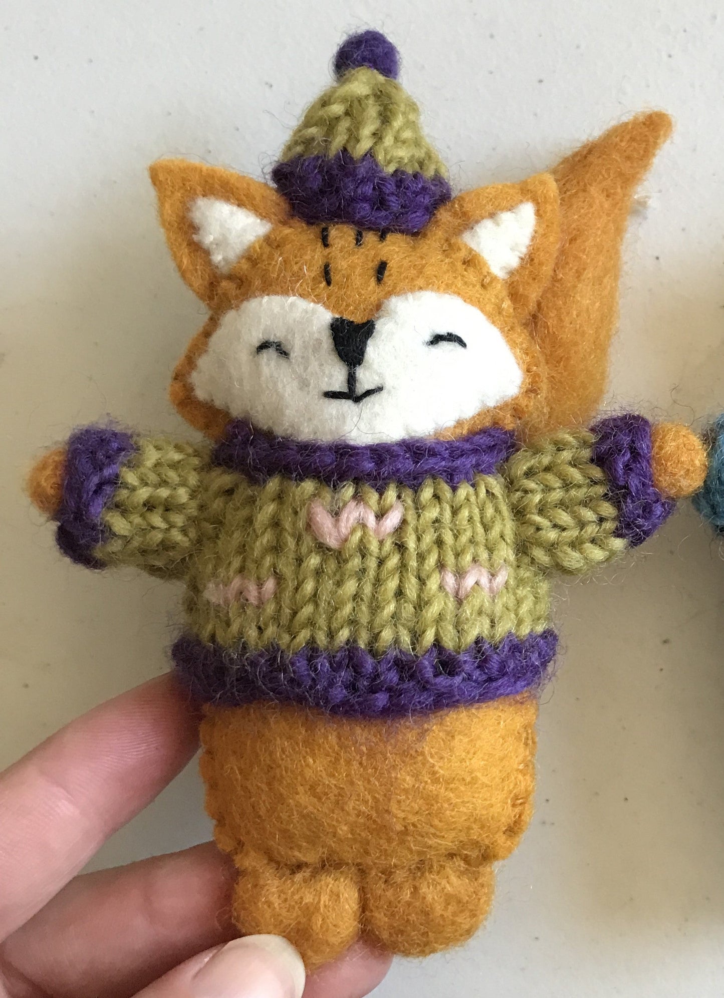 Close up of a felt ornament of a fox in a knitted yellow sweater and hat with purple trim and pink zig zags on it.