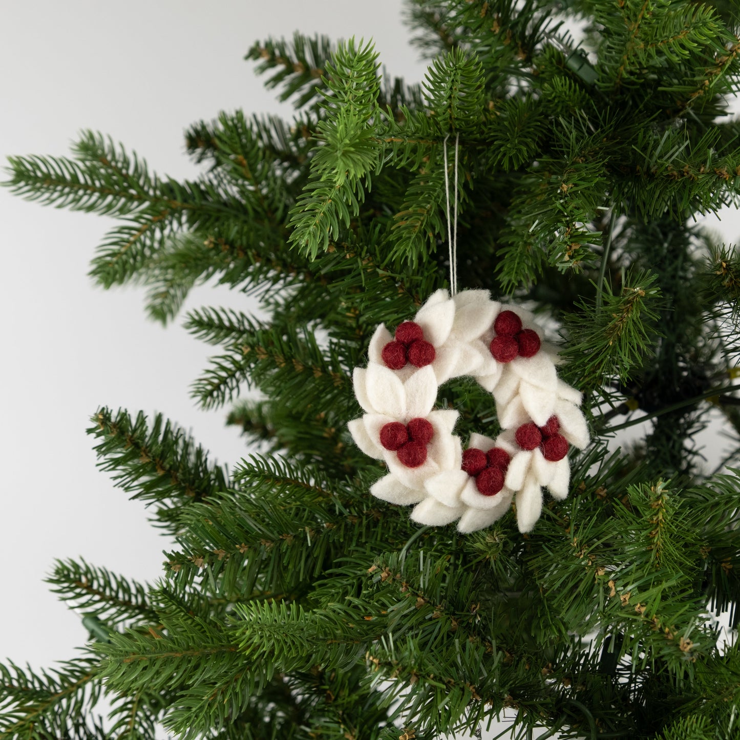 Felt ornament of a mini wreath in white with red berries hanging on a Christmas tree.