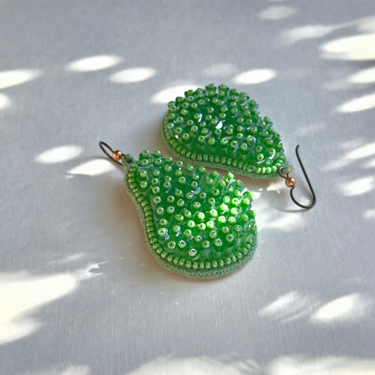 Pair of pincushion beaded green glass tassel earrings on shadowy background.