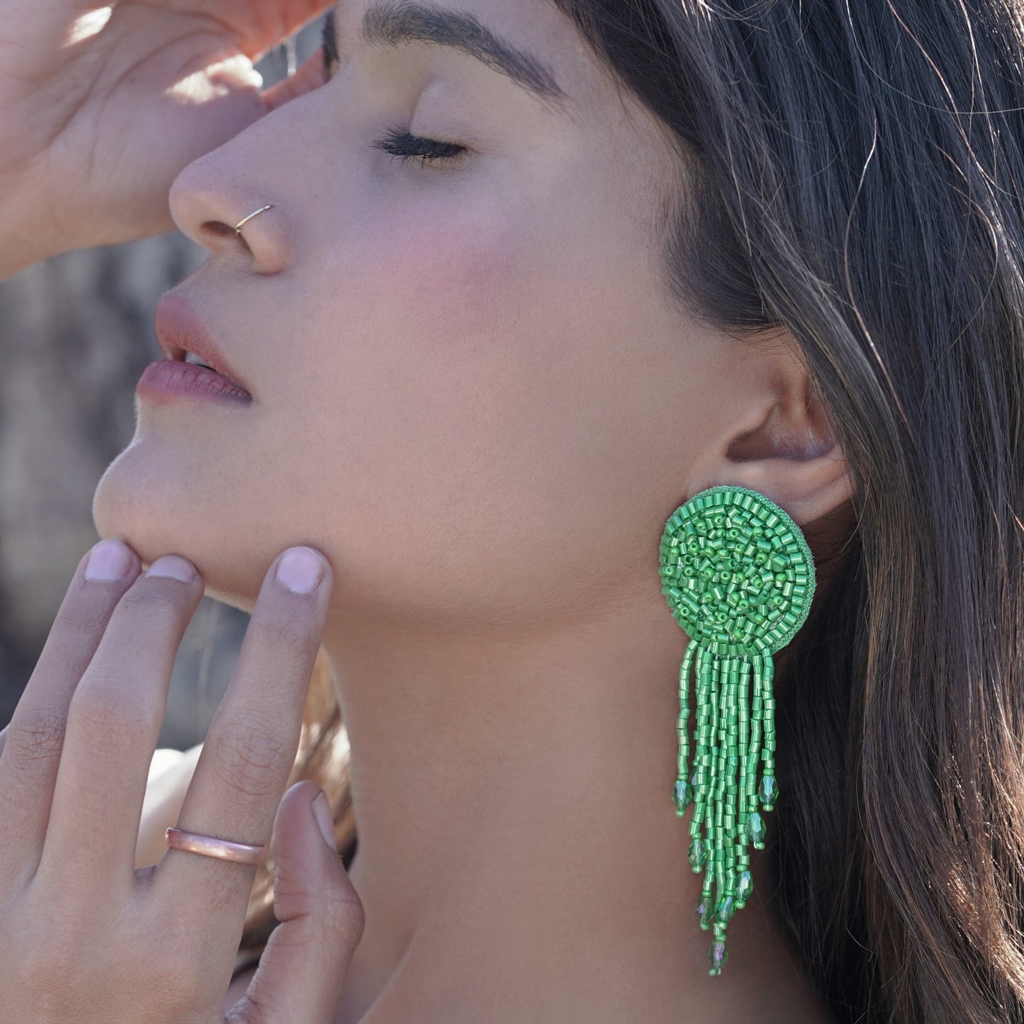 Left front view of woman with hands on face, wearing pair of green glass beaded tassel earrings.