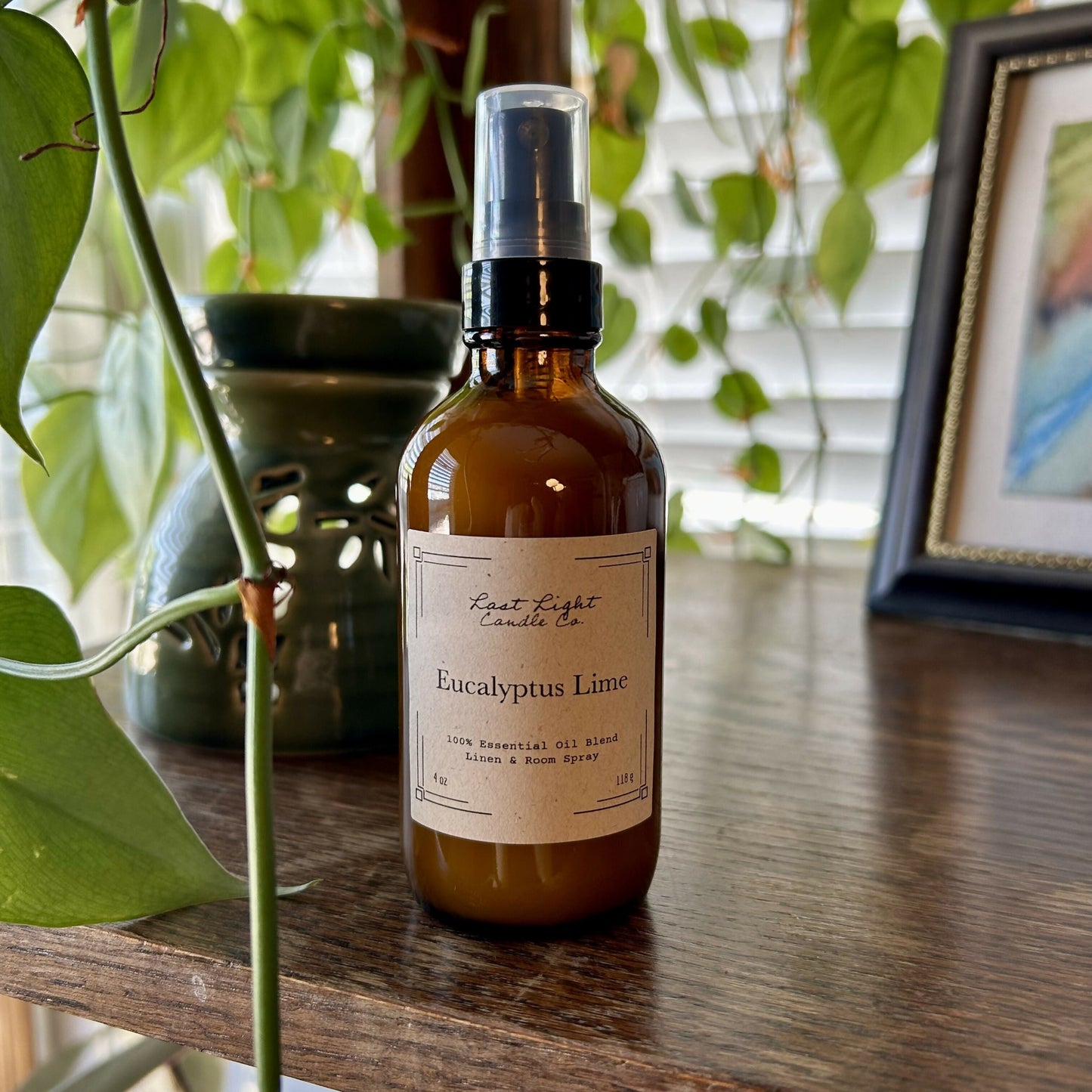 New Izela Eucalyptus Lime Room Spray Primary Photo. Amber jar with spray top on wood table next to hanging plant.