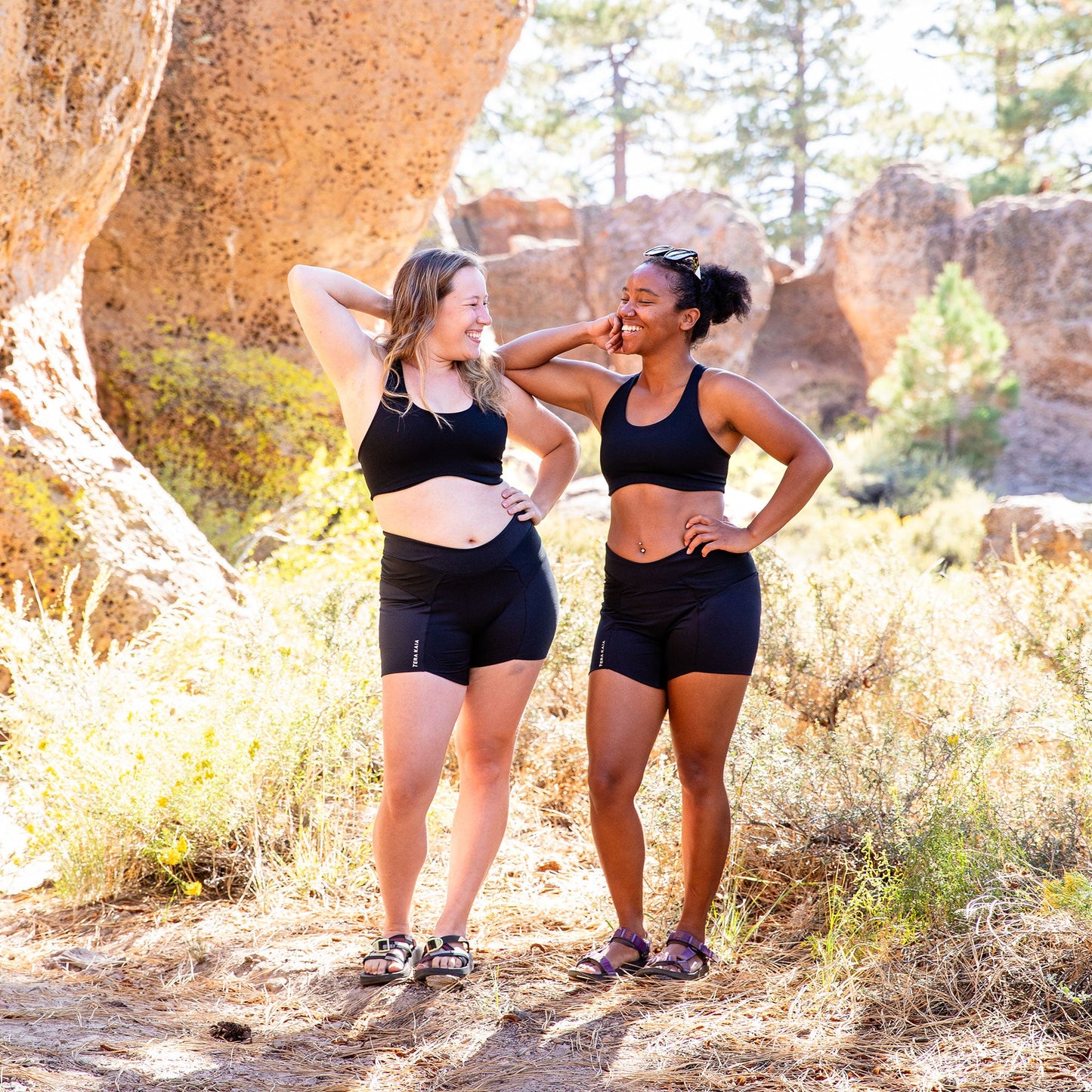 Two women near a rock, standing together with right elbows bent, each wearing black high cut top.