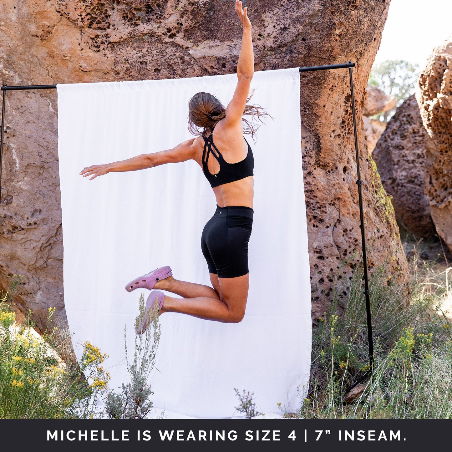 Back side view of woman jumping in front of a white sheet, hands in the air, wearing black toura short with 7" inseam.