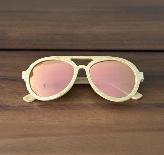 Avion style natural colored bamboo sunglasses with rose gold mirrored lenses folded sitting on dark brown wood.
