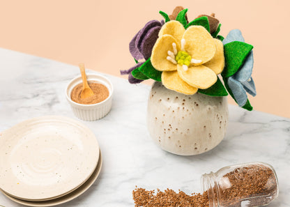 Purple, yellow, and blue felt Hepatica flowers in a tan round vase on table with plates.