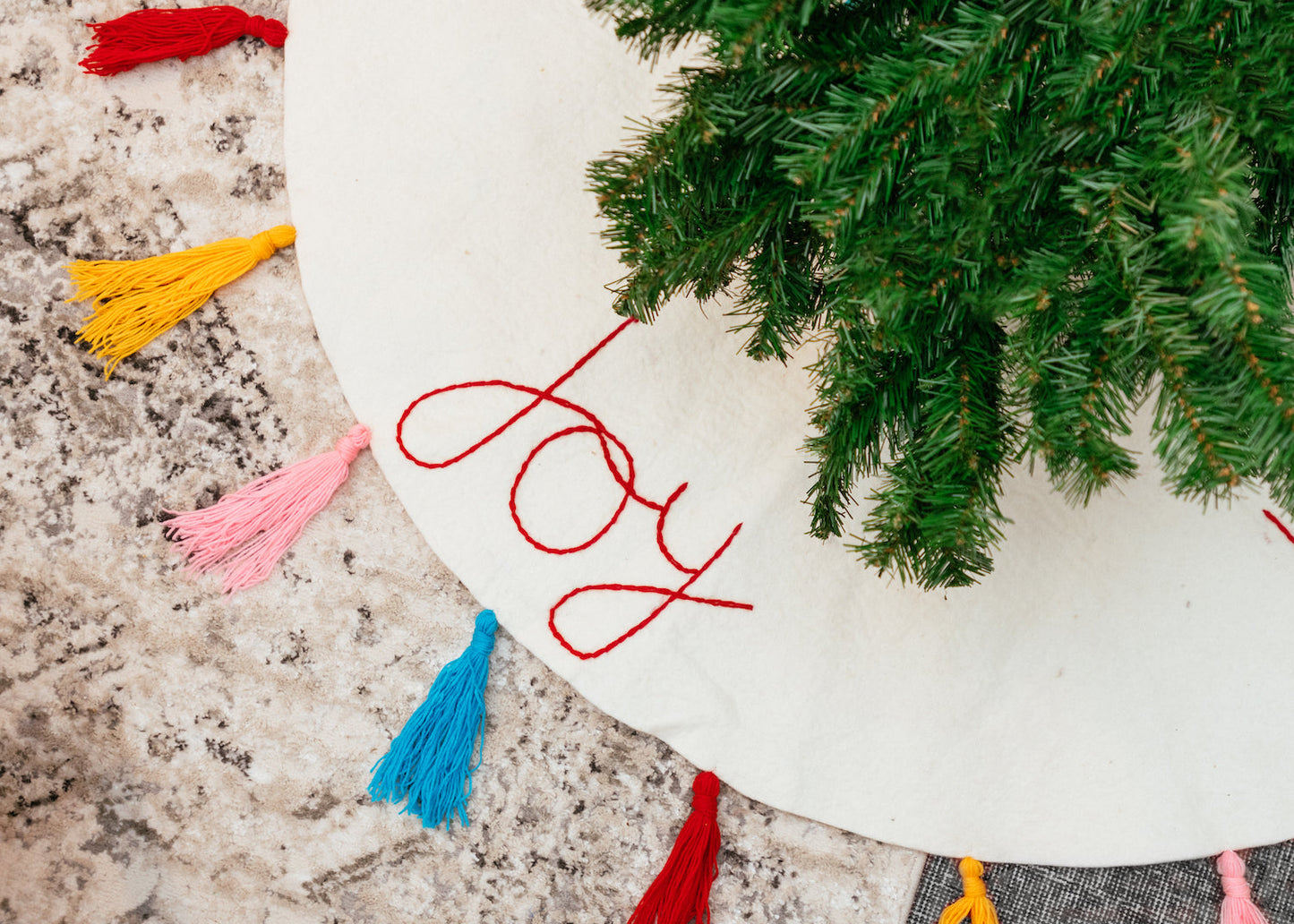 A white felt Christmas tree skirt with multicolored tassels around the edges and the words "peace" "love" and 'joy' embroidered around a tree.