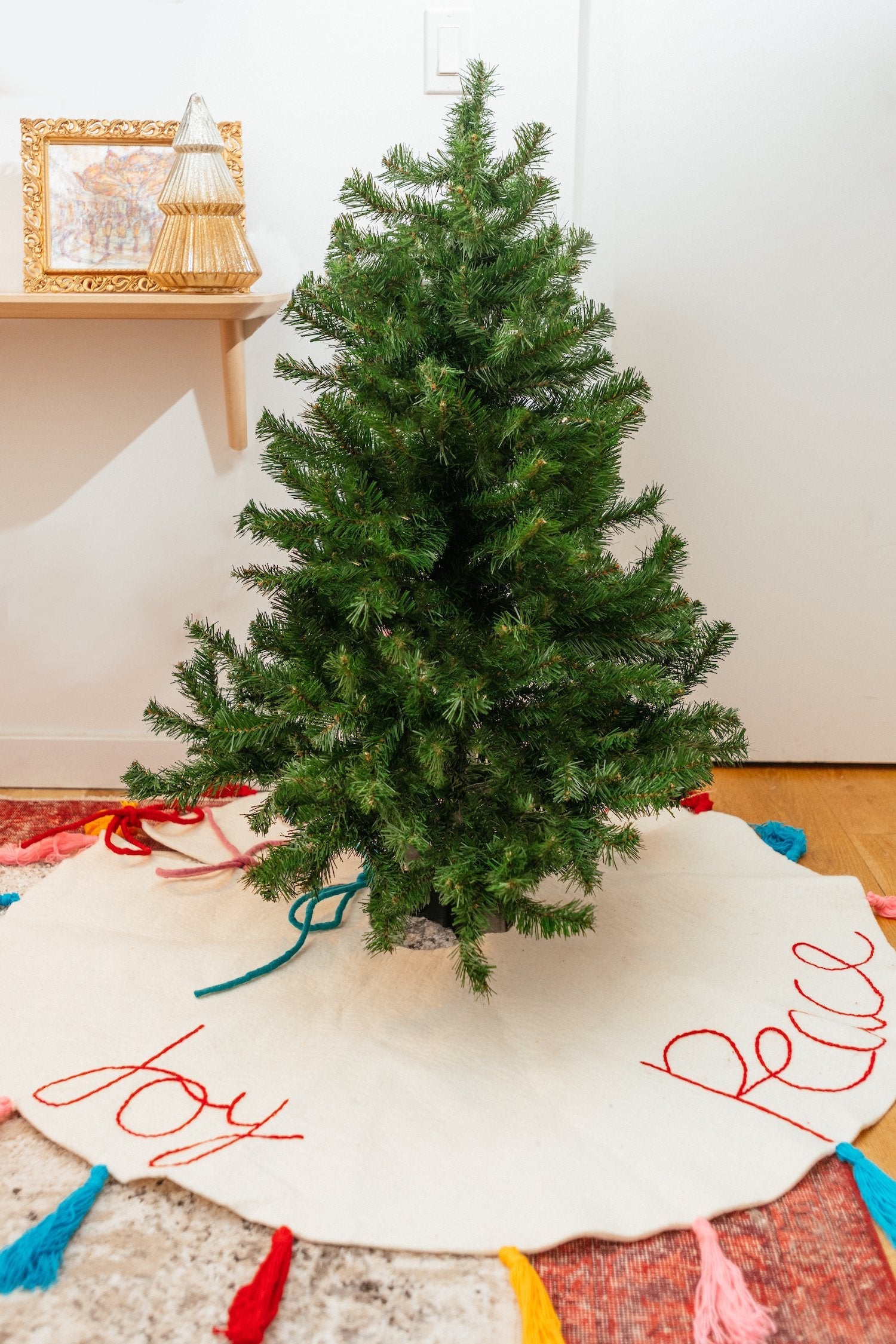 A white felt Christmas tree skirt with multicolored tassels around the edges and the words "peace" "love" and 'joy' embroidered around a tree.