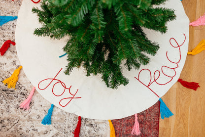 A white felt Christmas tree skirt with multicolored tassels around the edges and the words "peace" "love" and 'joy' embroidered around a tree.