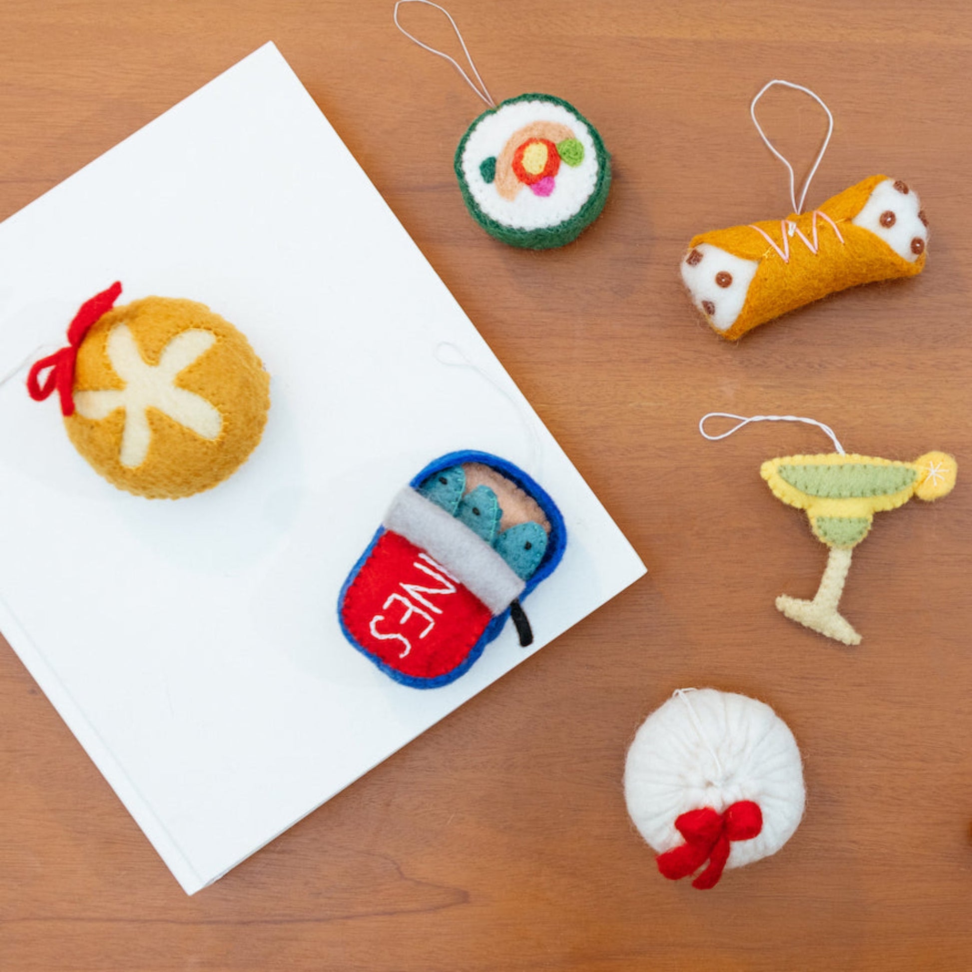 Scattered felt ornaments of a dumpling, a cannoli, sourdough bread, sardines, and a margarita.