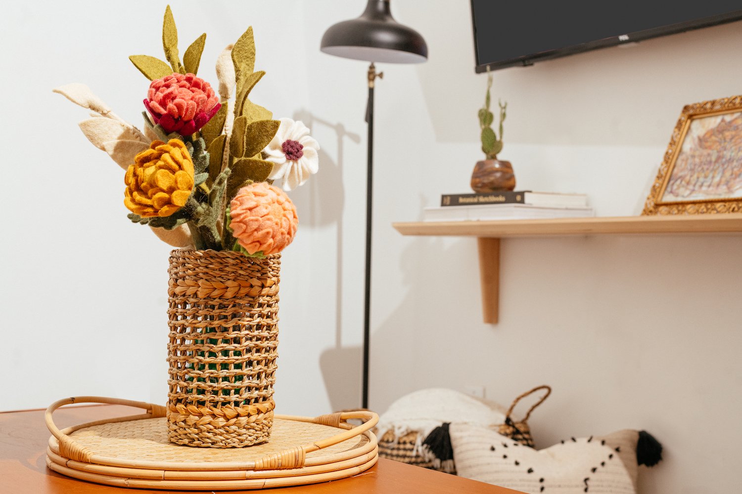 Tall brown woven vase with glass insert and felt flowers on brown table. Lamp and shelf in background..