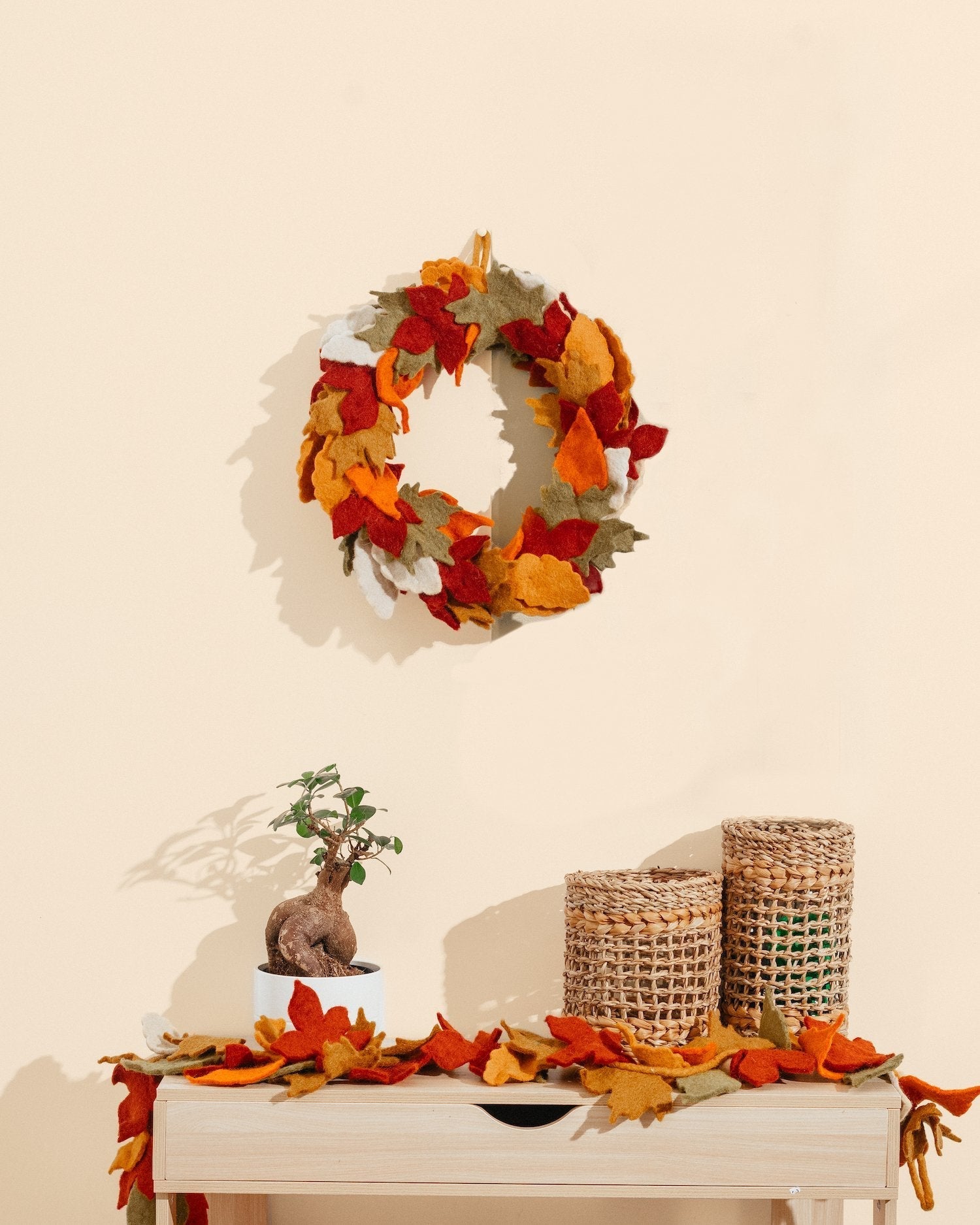 Short and tall brown woven vase with glass inserts on white table with wreath above on wall.