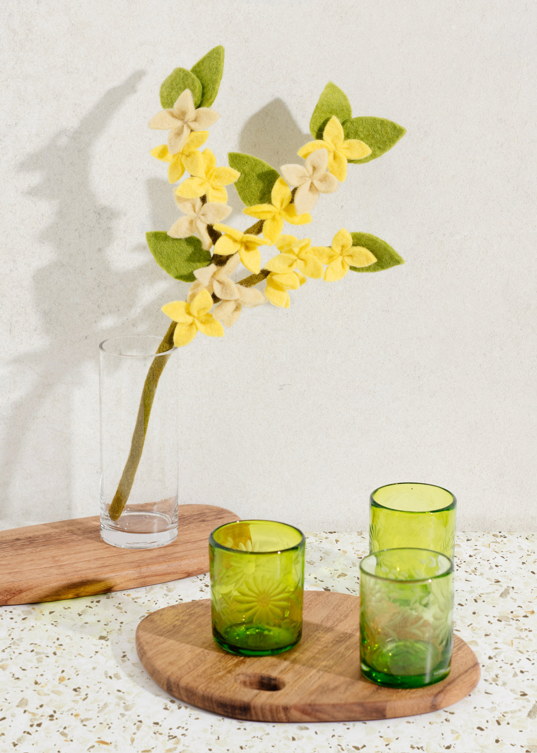 Brown wood oval serving board with yellow flowers on table with round serving board and green glasses in front.