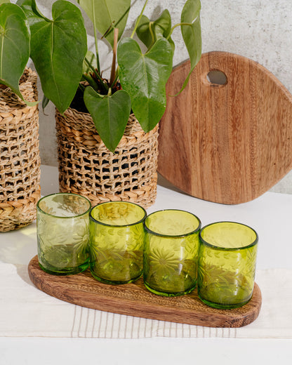 Brown wood round cutting board against wall, next to plants and additional long cutting board with four green glasses.