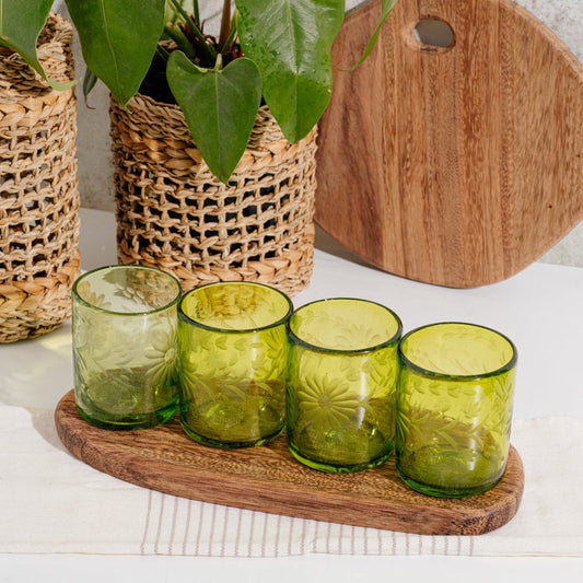 Brown wood oval serving board with four green glasses.  Additional items in background.