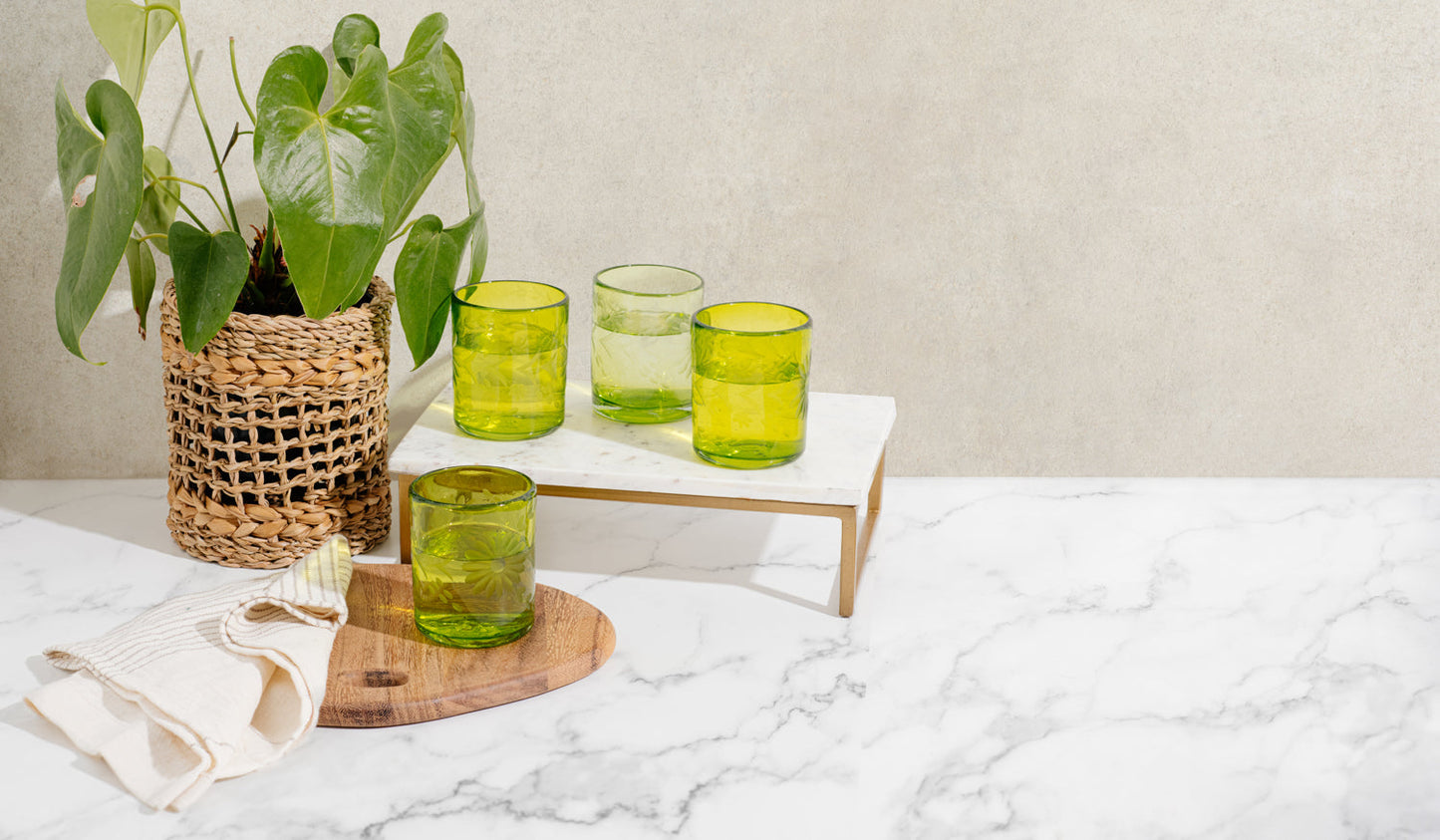 Edge of brown wood round cutting board with green glass next to towel, additional green glasses on tray and plant in woven vase..