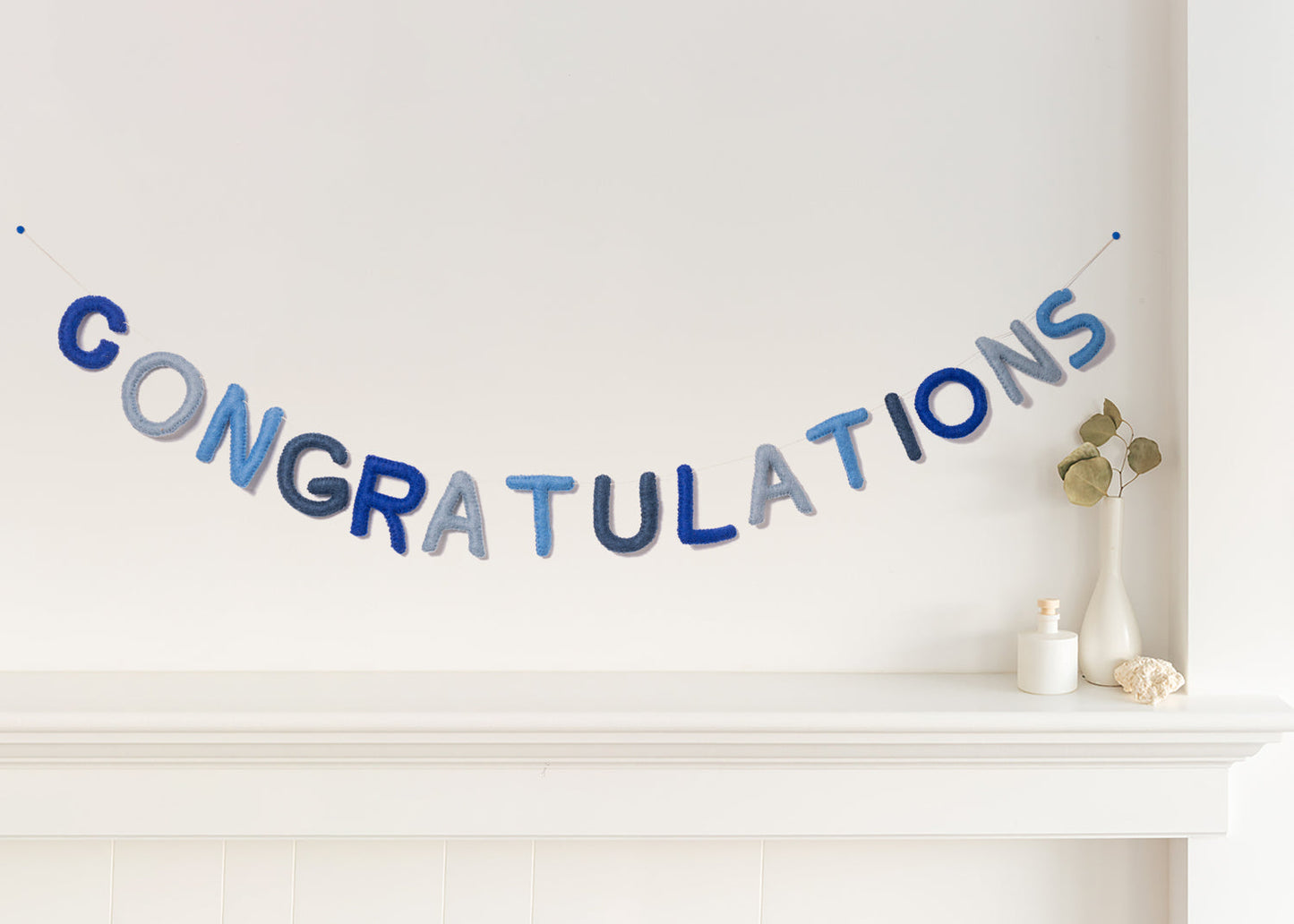 Felt garland spelling out Congratulations in various shades of blue alternating cobalt blue, pale blue, baby blue, and greyish blue hanging over a white mantel.