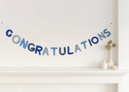 Felt garland spelling out Congratulations in various shades of blue alternating cobalt blue, pale blue, baby blue, and greyish blue hanging over a white mantel.
