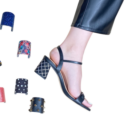 Woman's right foot and leg wearing black pants and black sandal with silver accents across toe strap and on heel. Multiple heel clips are shown, including blue, floral, silver diamond pattern, silver dot pattern, red, black with metal.