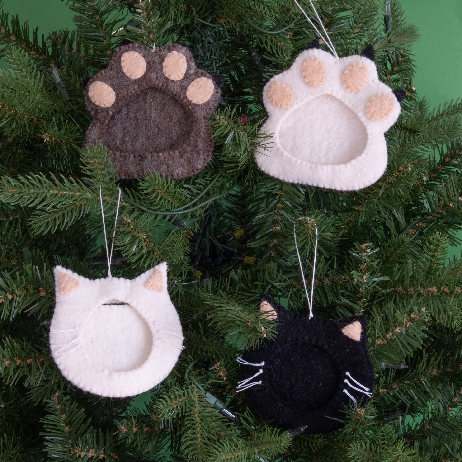 Felt ornaments of one black and one white cat face with a frame in the middle to put a photograph hanging on a Christmas tree with pawprint ornaments.