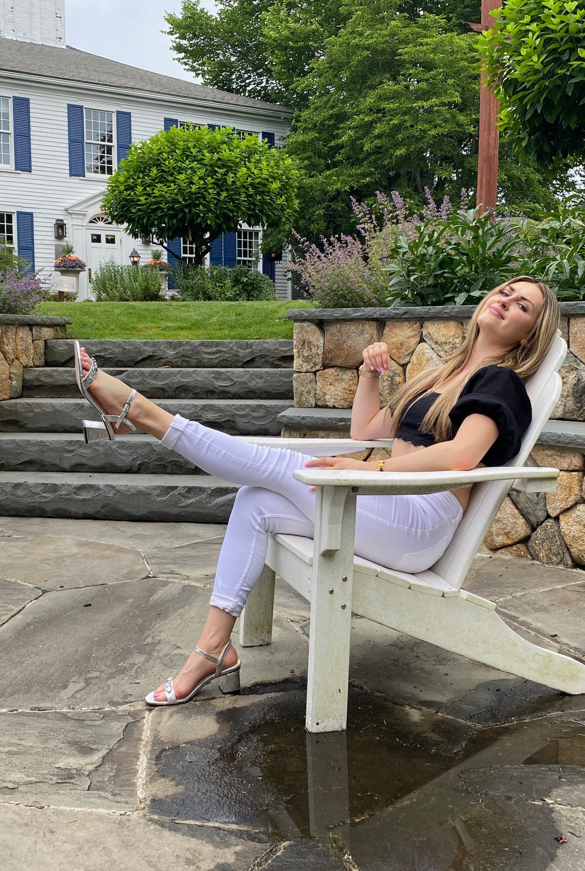 Woman sitting in chair, left leg in the air, wearing silver sandal.  Steps and house in background.