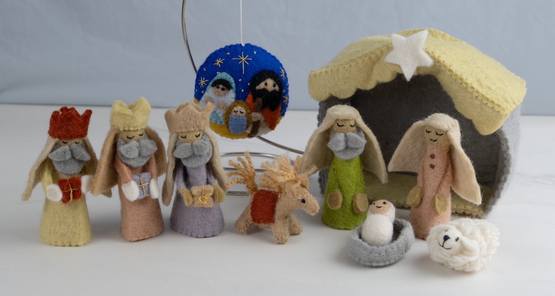 Felt ornament in a circle shape showing the nativity scene of Mary, Joseph, and baby Jesus underneath the night sky behind a full nativity scene set.